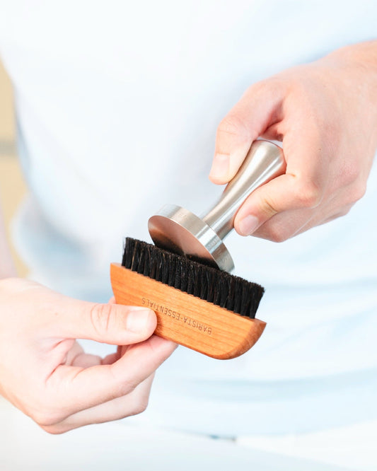 Image of a person holding a wooden brush and cleaning a stainless steel coffee tamper with it.
